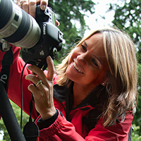 Smiling woman in a red jacket using a DSLR camera outdoors, adjusting the lens.
